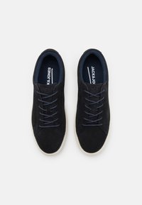 Adidași negri din suede cu șireturi de un albastru închis și o talpă din cauciuc alb contrastant. Marca este vizibilă pe tălpi. Design simplu, de profil joas.