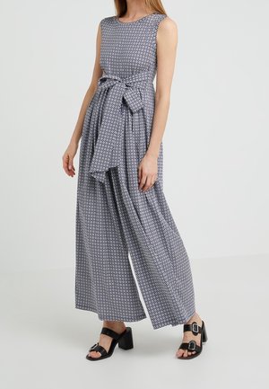 Kvinde iført ærmeløs buksedragt med vide ben, lille geometrisk mønster og stor sløjfe i taljen, kombineret med sorte hælsandaler med spænde.