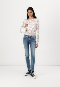 Hosszú ujjú fehér virágmintás blúz, sötétkék skinny farmernadrág és fekete magas szárú sportcipők. Kiegészítő: kis fehér kézitáska. Egyszerű, modern dizájn.
