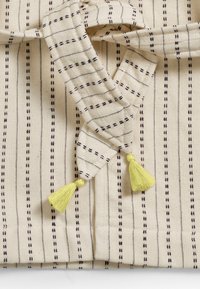 Puuvillakangas kreemitaustaga, kitsaste mustade triipudega ja kollaste pompoonidega otstes, millel on keeruline kudumise tekstuur.