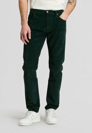 Homme portant un pantalon en velours côtelé vert foncé et des baskets blanches, debout avec une main dans la poche devant un fond clair uni.