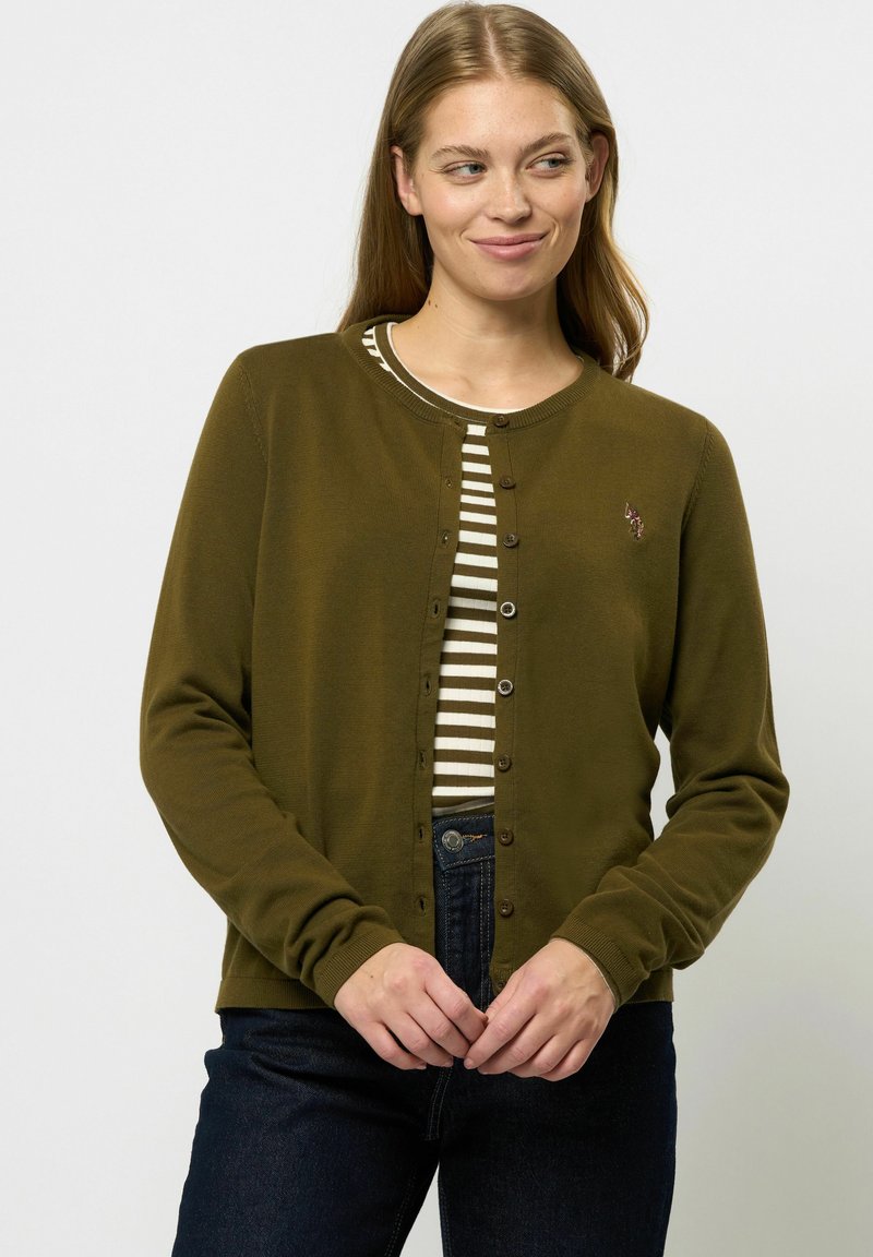 Olivgrön kofta med långa ärmar och knappstängning, över en krämfärgad och mörk randig topp. På vänster sida finns en anmärkningsvärd broderad detalj.