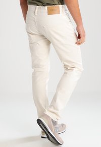 Pantalones de mezclilla blancos con un corte recto, que cuentan con dos bolsillos traseros y una etiqueta de cuero marrón en la cintura. Combinados con zapatillas deportivas grises.