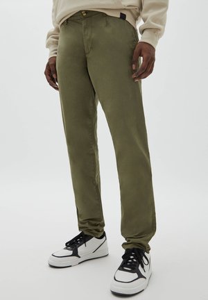 Homme portant un pantalon vert olive, un sweat-shirt beige et des baskets blanches avec des détails noirs, debout sur un fond uni.