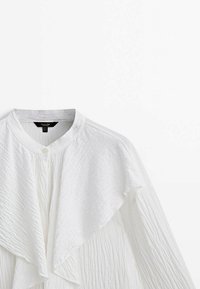 Chemise blanche boutonnée avec un tissu texturé froissé, dotée d'un devant à volants, d'un col montant et de manches longues.
