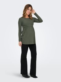 Tehotná žena v olivovo zelenej dlhorukávovej blúzke a čiernych nohaviciach stojaca, jedna ruka jej siaha na vlasy, na jednoduchej pozadí.