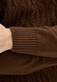 Maglione marrone a fantasia treccia con polsini a costine, in primo piano per mostrare i dettagli di texture e colore della manica e del braccio.