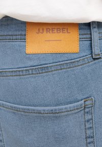 Poche de jean en denim bleu clair avec une étiquette en cuir marron cousue au-dessus, embossée avec "JJ REBEL" en lettres majuscules.