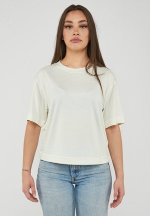 Giovane donna con lunghi capelli castani che indossa una maglietta bianca a maniche corte e larga e jeans azzurri chiari, in piedi su uno sfondo semplice.