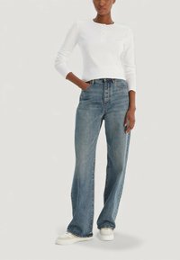 Bílý top henley s dlouhým rukávem a zapínáním na knoflíky, doplněný širokými modrými džínami. Model nosí bílé tenisky; pohodlný střih a volná silueta.