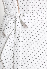 Robe en tissu blanc à motif de pois noirs, avec une ceinture à nouer à la taille et une fermeture éclair visible à l'avant. Texture légère.