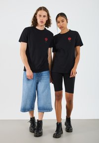 Černé bavlněné trička s výšivkou červené růže, nošená s modrými denimo culottes a černými cyklistickými šortkami. Oba mají na sobě černé šněrovací boty.