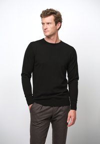 Schwarzer, langärmeliger Pullover aus weichem Stoff mit Rundhalsausschnitt, kombiniert mit grauen, strukturierten Hosen, Hände in den Taschen, neutraler Hintergrund.