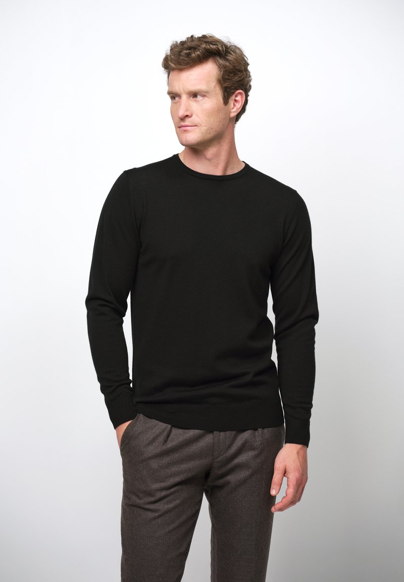 Schwarzer, langärmeliger Pullover aus weichem Stoff mit Rundhalsausschnitt, kombiniert mit grauen, strukturierten Hosen, Hände in den Taschen, neutraler Hintergrund.