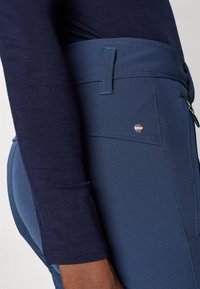 Marineblå stretchbukser med en glat overflade, der har en lynlåslomme og en flad talje, accentueret af en lille metallisk logo detalje.