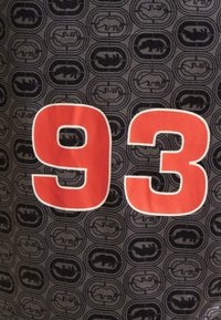 Svart stoff med et gjentatt sirkulært mønster med flaggermus-logoer. Stor rød "93" som er vist tydelig i midten.