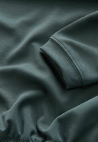Tissu vert foncé doux avec coutures visibles et poignet élastique, montrant la texture détaillée et les plis d'une manche de vêtement.