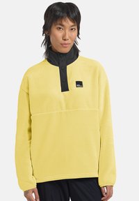 Pullover in pile giallo con colletto a metà zip nera, bottoni a pressione e una piccola etichetta con logo sul davanti. Tessuto morbido e vestibilità comoda.