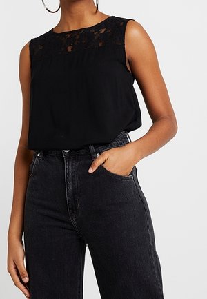 Blouse noire sans manches avec détail en dentelle au niveau de l'encolure, associée à un jean noir taille haute, une main dans la poche.