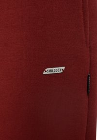 Rød strikket stof med en blød tekstur. Har et lille "SMILODOX" logo mærke syet på, med en kontrasterende syning detalje og en sidelomme.