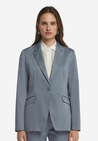 Ljusblå kavaj med en enkel knappstängning, spetsiga rever och två framfickor. Bärs över en vit skjorta med krage.