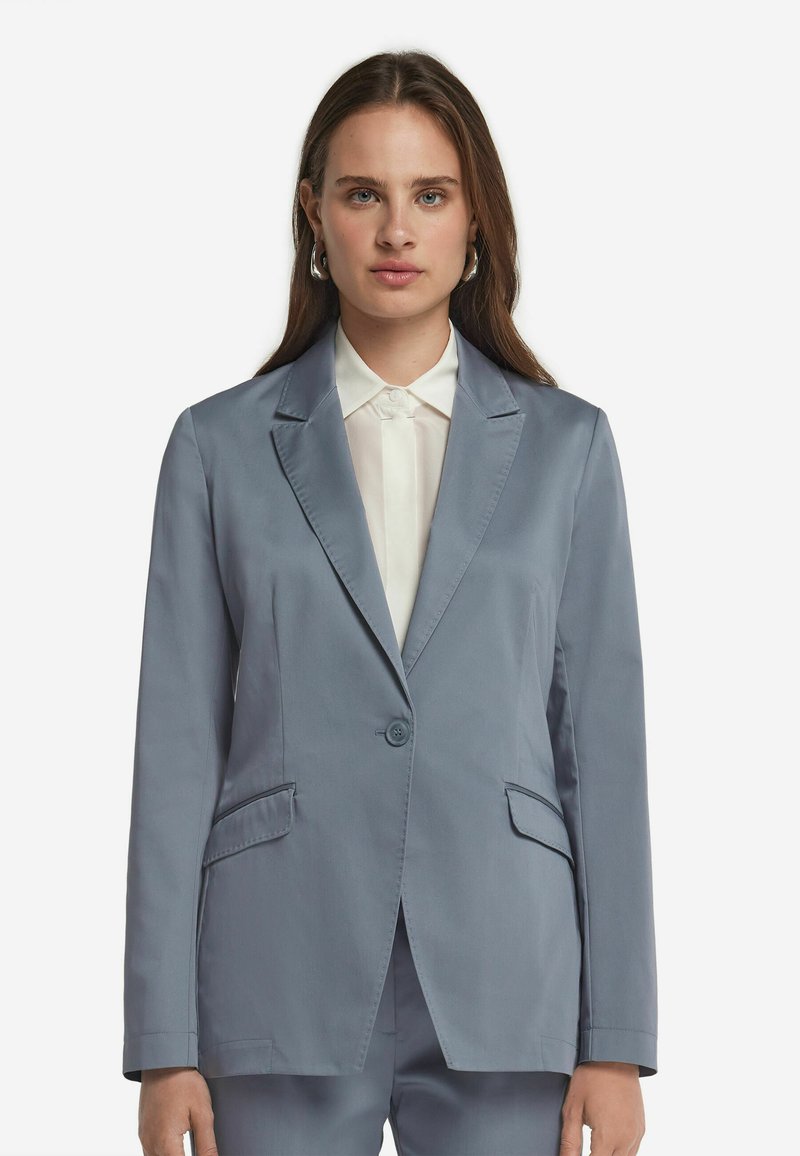 Ljusblå kavaj med en enkel knappstängning, spetsiga rever och två framfickor. Bärs över en vit skjorta med krage.