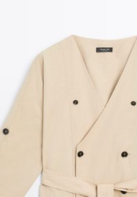 Beige Wickelkleid mit tiefem V-Ausschnitt, doppelten schwarzen Knopf-Details und Bindegürtel. Aus glattem Stoff und mit dezenten Nähten.