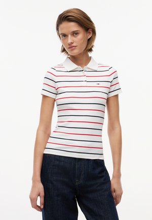 Kobieta ubrana w białą koszulkę polo z czerwonymi i granatowymi poziomymi paskami oraz ciemnoniebieskie dżinsy, stojąca na jednolitym białym tle.