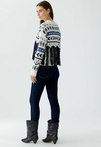 Gebreide trui met marineblauwe en witte geometrische patronen, franjes aan de onderkant, gecombineerd met donkerblauwe jeans en zwarte kniehoge laarzen met een spitse neus.