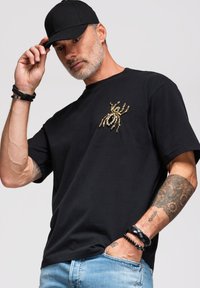 Homme avec casquette noire et t-shirt noir avec design d'araignée dorée, portant des bracelets, montrant un avant-bras tatoué, une main dans la poche du jean.