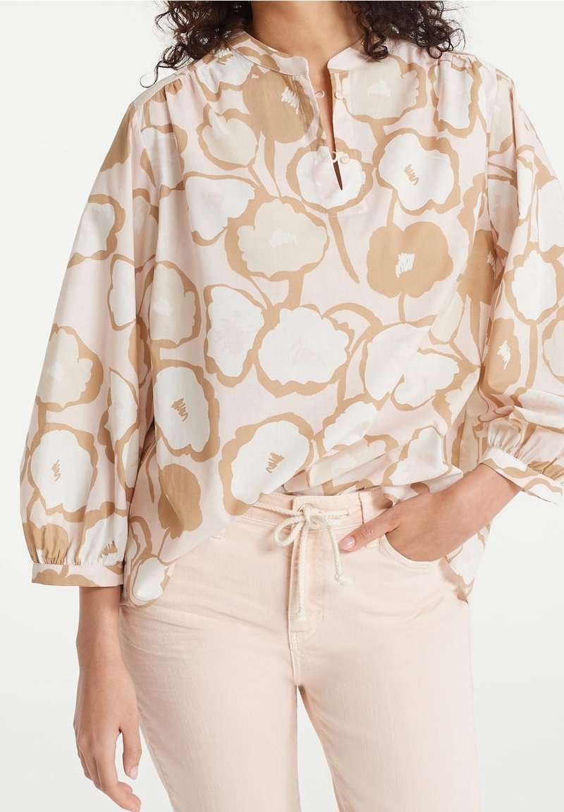 Weit geschnittene Bluse in Hellrosa mit einem floralen Muster in Beige und Weiß. Sie hat einen kleinen Schlitz am Ausschnitt und elastische Ärmel.