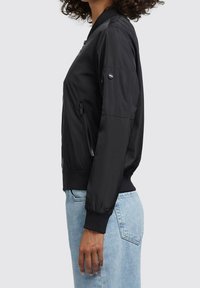 Černá bomber bunda s hladkou, lesklou texturou, zipovými bočními kapsami, žebrovanými manžetami a přiléhavým pasem.