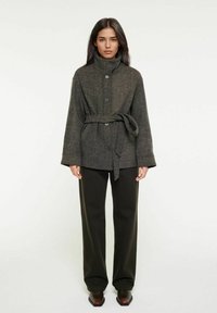 Grå texturerad ullkappa med hög krage, knappar framtill och knytbart midjebälte, i kombination med mörka byxor och skor med spetsig tå.