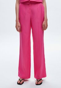 Roze wijde broek van soepel materiaal met centrale plooien aan de voorkant. Gestyleerd met zwarte slippers die zichtbaar zijn bij de enkel.