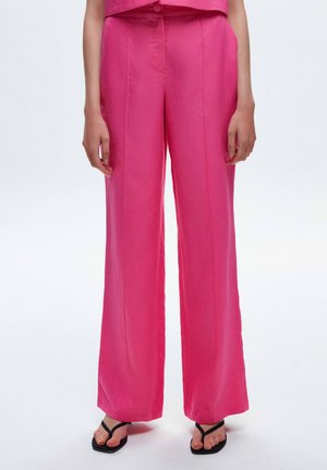 Rosa weitgeschnittene Hose aus glattem Stoff mit mittigen Falten. Kombiniert mit schwarzen Flip-Flops, die am Knöchel sichtbar sind.