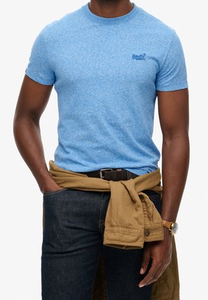 Man die een aangesloten lichtblauw t-shirt draagt, donkere spijkerbroek, bruine riem, beige jas in de taille gestrikt en een gouden horloge, poserend met één hand in de zak.