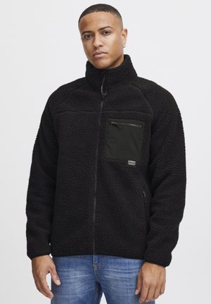 Black fleece jacket with high collar, zippered chest pocket, and side pockets. Features a textured, soft material and contrasting pocket fabric.