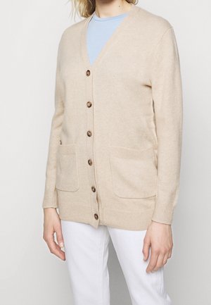 Femme portant un cardigan beige boutonné avec deux poches avant par-dessus une chemise bleu clair et un pantalon blanc, debout devant un fond uni.
