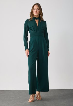 Green jumpsuit with long sleeves, keyhole neckline, pleated bodice, wide-leg design, made from stretchy fabric, paired with gold sandals.