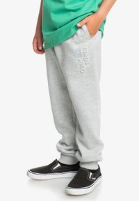 Pantalones de chándal grises hechos de una mezcla de algodón, con bolsillos laterales, puños elásticos y un logotipo en relieve en azul claro. Zapatillas negras de tipo slip-on.