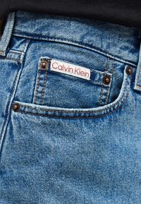 Τσέπη μπλε τζιν με χαλκομανίες και λευκή ετικέτα ραμμένη στην τσέπη, που εμφανίζει το «Calvin Klein» με κόκκινο κείμενο.