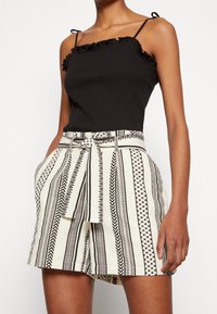 Débardeur noir côtelé avec des bords à volants, associé à un short taille haute crème présentant des motifs rayés géométriques noirs et une ceinture nouée.