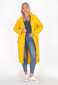 Sárga vízálló esőkabát kapucnival, patent gombokkal és oldalsó zsebekkel, csíkos póló és kék farmert viselve.