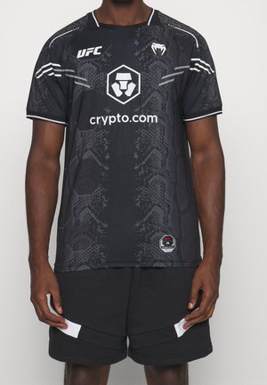 Mężczyzna noszący czarną sportową koszulkę UFC z wzorem węża, logo crypto.com oraz czarne spodenki z białymi akcentami, stojący na jednolitym tle.