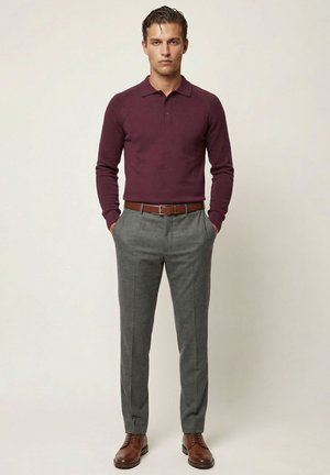 Mężczyzna stojący z rękami w kieszeniach, ubrany w bordową koszulkę polo z długim rękawem, szare spodnie, brązowy pasek i brązowe eleganckie buty.