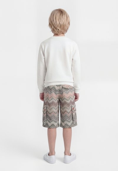 Enfant blond vu de dos, portant une chemise blanche à manches longues, un short cargo à motifs et des baskets blanches sur un fond uni.