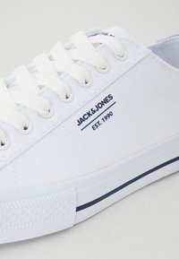 Białe płótno trampki z okrągłym czubkiem, płaskimi białymi sznurowadłami, granatowym logo "JACK & JONES EST. 1990" oraz granatowym wykończeniem wzdłuż podeszwy.