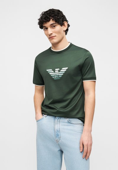 Νεαρός άνδρας με σγουρά μαλλιά που φοράει σκούρο πράσινο κοντομάνικο πουκάμισο με λευκή επένδυση και λογότυπο αετού με ρίγες, σε συνδυασμό με ανοιχτόχρωμο μπλε τζιν.