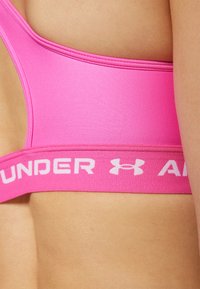 Rozā sporta krūšturis ar gludu tekstūru un platām siksnām, kurās ir balti burti "UNDER ARMOUR". Dizains nodrošina ērtu piegulšanu.