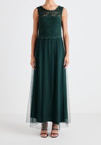 Pitkä, tummanvihreä mekko, jossa on pitsipaita, jossa on aallotettu reuna ja virtaava, läpikuultava hame. Käytetty mustien hihnatossujen kanssa.